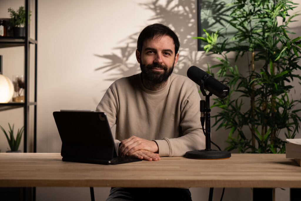 Homme barbu en sweat beige assis à un bureau, face caméra, prêt à enregistrer avec un micro podcast.