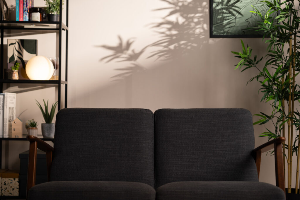 Détail du décor L’Essentiel avec canapé gris en avant-plan et ombres nettes de feuilles projetées sur le mur, créant une ambiance chaleureuse.