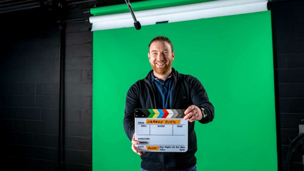 tournage-fond-vert-slate-bts Homme souriant tenant un clap de cinéma devant un fond vert.