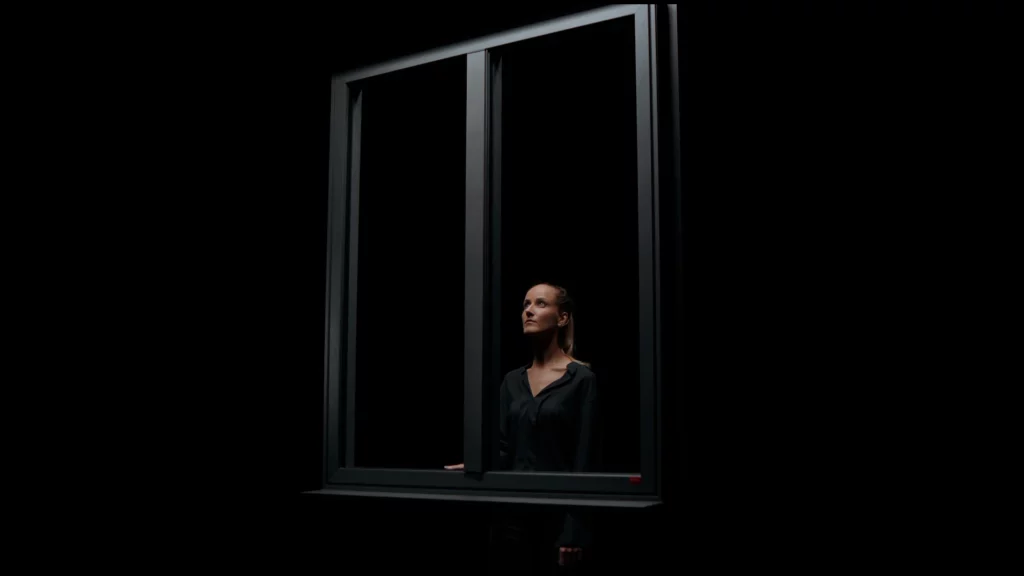 bochassy-fenetre-premium-shooting-pub-cinematique-studio-401-02 Photographie publicitaire pour la campagne "Fenêtre Premium" de Bochassy, réalisée au Studio 401 à Rennes. Une femme regarde vers le haut, cadrée à travers le châssis d'une fenêtre en aluminium noir. La scène est plongée dans l'obscurité totale du plateau "boîte noire", avec une lumière latérale précise qui vient sculpter les traits du visage et souligner la finition mate du produit. Ce visuel illustre le savoir-faire du studio pour des packshots produits mis en scène avec une esthétique haut de gamme et épurée.