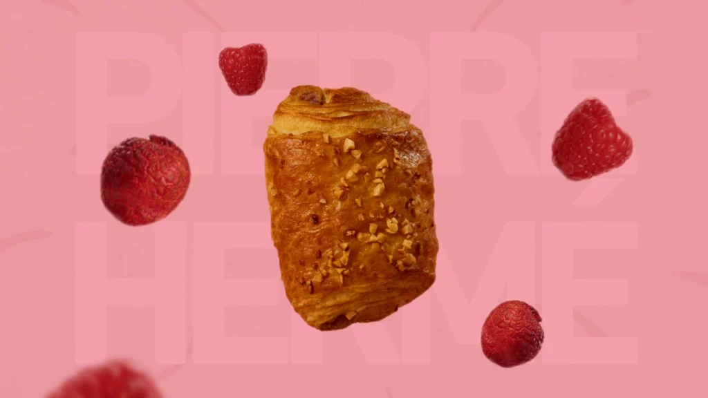 bridor-pierre-herme-signature-framboise-still-life-studio-401-03 Photographie culinaire publicitaire pour la collaboration "Bridor x Pierre Hermé", réalisée au Studio 401 à Rennes. Une viennoiserie feuilletée dorée "Signature Framboise" lévite au centre de l'image, entourée de framboises fraîches et de litchis en suspension. Le fond est d'un rose uni avec le nom "PIERRE HERMÉ" inscrit en filigrane large et discret. Ce visuel dynamique et graphique met en avant l'aspect créatif et premium de la gamme d'agrumes et de fruits rouges produite en studio.