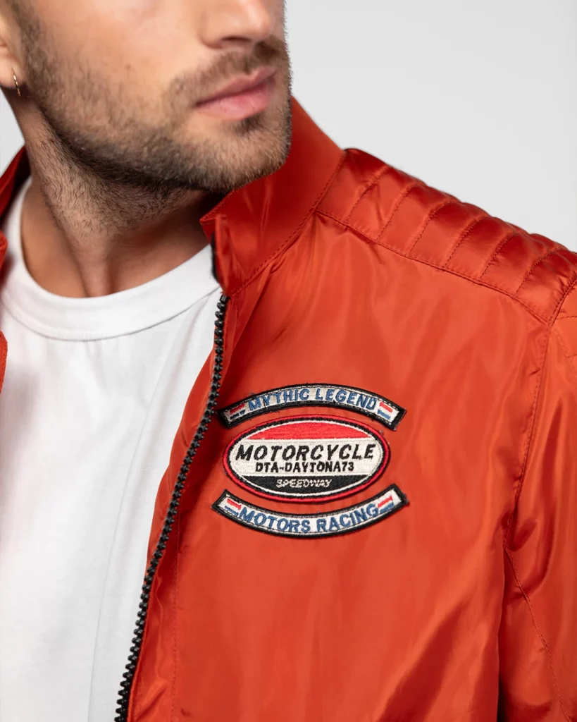 daytona-73-detail-blouson-orange-shooting-photo-studio-401 Photographie publicitaire en gros plan (close-up) montrant les détails d'un blouson orange de la marque Daytona 73, réalisée au Studio 401 à Rennes. Le visuel met en avant la texture du tissu satiné et les écussons brodés de style vintage ("Mythic Legend", "Motorcycle"). L'éclairage de studio précis souligne la qualité des finitions et les reflets du textile pour un catalogue de mode haut de gamme.