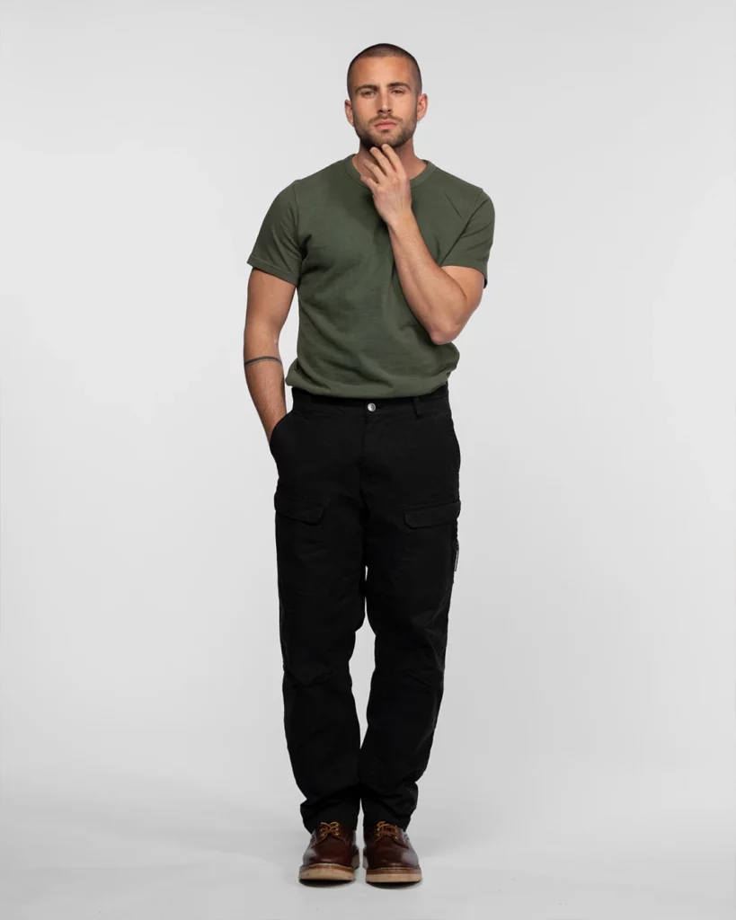 daytona-73-shooting-photo-homme-e-commerce-studio-401 Photographie de mode e-commerce pour la marque Daytona 73, réalisée au Studio 401 à Rennes. Un modèle masculin pose en pied devant un fond cyclorama gris clair uniforme. Il porte un t-shirt vert kaki, un pantalon cargo noir et des chaussures en cuir marron. L'éclairage studio est parfaitement équilibré pour mettre en valeur les textures et les coupes des vêtements. Ce visuel démontre la capacité du plateau à accueillir des productions de catalogue à fort volume avec un rendu professionnel constant.