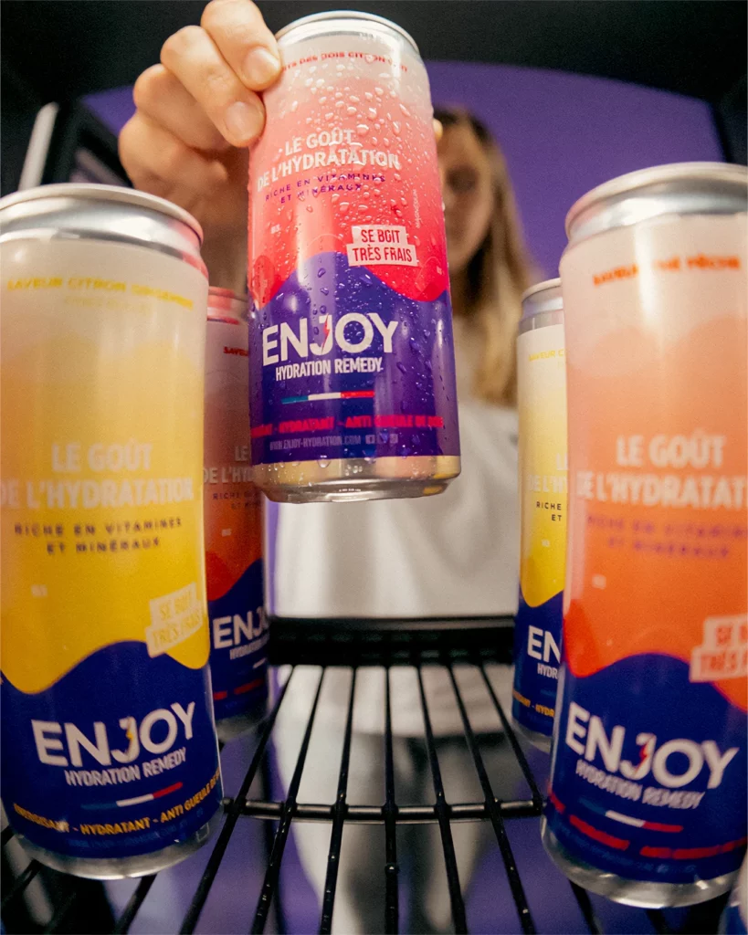 enjoy-hydratation-shooting-lifestyle-pov-frigo-studio-401 Photographie publicitaire créative pour "Enjoy Hydratation", réalisée au Studio 401 à Rennes. Le visuel adopte un point de vue immersif depuis l'intérieur d'un réfrigérateur. Une main saisit une canette de boisson fraîche et givrée (saveur Fruits des bois) au centre, tandis que d'autres canettes sont disposées au premier plan sur la grille. Une silhouette est visible en arrière-plan sur un fond violet. Cette image illustre l'expertise du studio pour créer des contenus social media originaux et engageants.