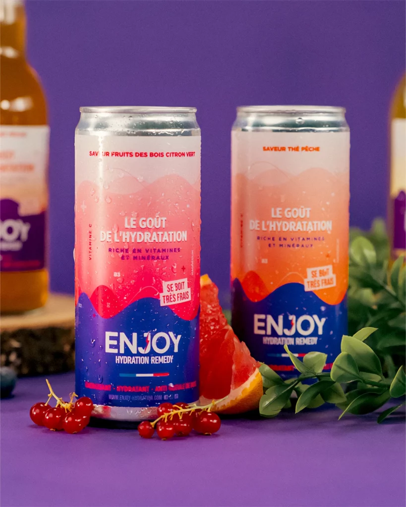 enjoy-hydratation-shooting-produit-canette-fraicheur-studio-401 Photographie de produit publicitaire pour les boissons "Enjoy Hydratation", réalisée au Studio 401 à Rennes. Deux canettes de "Hydration Remedy" (saveurs Fruits des bois et Thé pêche) sont mises en scène sur un fond violet vibrant. Les canettes sont couvertes de gouttelettes de condensation pour accentuer l'aspect rafraîchissant. Le décor est complété par un quartier de pamplemousse, des grappes de groseilles rouges et du feuillage vert. L'éclairage studio met en valeur les couleurs vives et le design dynamique du packaging.