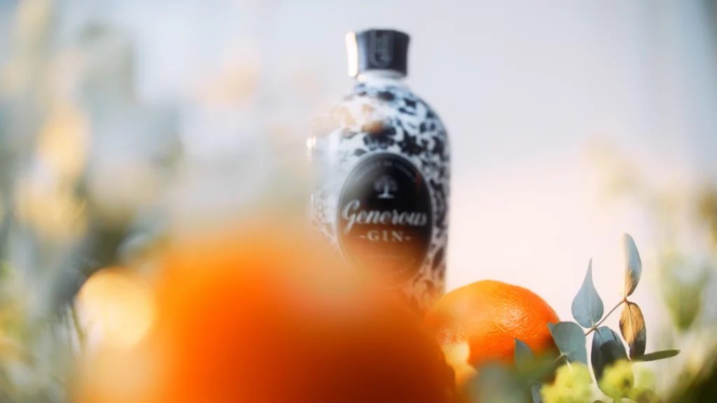 generous-gin-packshot-lifestyle-bokeh-lumineux-studio-401 Photographie publicitaire pour la marque "Generous Gin" réalisée au Studio 401 à Rennes. La bouteille aux motifs floraux noirs est mise en scène dans un environnement lumineux et vaporeux. Un premier plan flou orangé crée un effet de bokeh chaleureux, tandis qu'une orange entière et des feuilles d'eucalyptus encadrent le produit. L'éclairage doux et solaire souligne l'aspect naturel et haut de gamme de la marque pour une communication spiritueux premium.