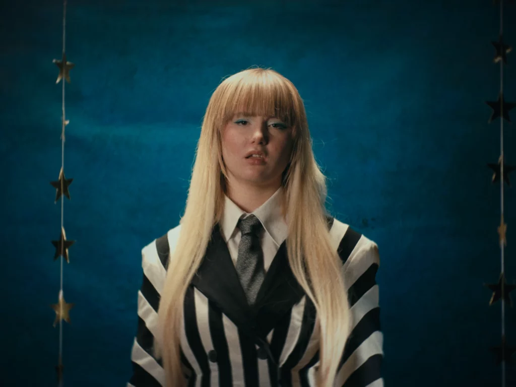 jeanne-bonjour-clip-finir-par-en-rire-decor-etoiles-studio-401 Jeanne Bonjour cadrée devant un fond bleu texturé avec des étoiles dorées suspendues, extrait du clip "Finir par en rire". La mise en scène minimaliste mais impactante souligne la polyvalence du Studio 401 pour créer des univers visuels uniques en intérieur.