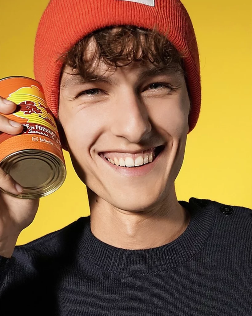 la-belle-iloise-chauds-les-coeurs-portrait-lifestyle-studio-401-01 Photographie publicitaire pour la campagne "Chauds les Cœurs" de La Belle-Iloise, réalisée au Studio 401 à Rennes. Un homme souriant, portant un bonnet orange vif et un pull en maille bleu marine, tient une conserve de soupe de poisson contre son visage. L'image utilise un fond jaune éclatant, créant un style "color block" dynamique. Ce portrait met en avant l'expertise du studio en photographie lifestyle et publicitaire pour le secteur agroalimentaire.