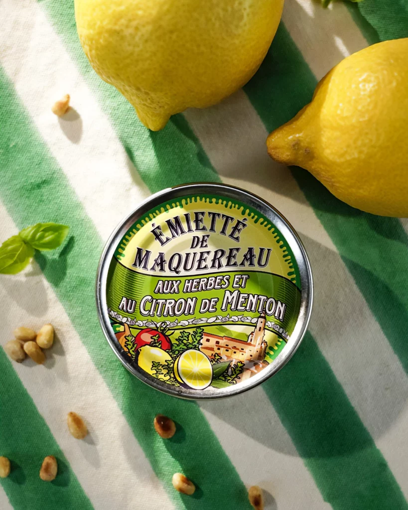 la-belle-iloise-maquereau-citron-menton-shooting-culinaire-studio-401 Photographie culinaire "flat lay" d'une boîte d'Emietté de Maquereau aux herbes et au citron de Menton de la marque La Belle-Iloise, réalisée au Studio 401 à Rennes. La boîte est centrée sur un linge à rayures vertes et blanches, entourée de deux citrons jaunes entiers, de pignons de pin éparpillés et d'une feuille de basilic. La lumière vive et contrastée simule un ensoleillement naturel, mettant en avant la fraîcheur et la qualité premium du produit.