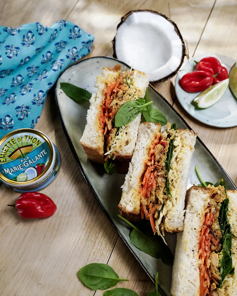 la-belle-iloise-recette-thon-marie-galante-shooting-culinaire-studio-401 Photographie culinaire en flat lay (vue de dessus) pour une recette de La Belle-Iloise, réalisée au Studio 401 à Rennes. Des sandwiches club gourmands garnis d'émietté de thon, carottes et épinards sont présentés sur une assiette en céramique. Le décor sur bois clair inclut une boîte d'Émietté de Thon à la Marie-Galante, une demi-noix de coco, des piments habaneros et du citron vert. La lumière chaude et directionnelle évoque une ambiance exotique et solaire, soulignant le savoir-faire du studio en stylisme culinaire.
