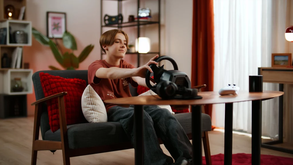 studio-tournage-rennes-set-design-gaming-lifestyle Jeune acteur utilisant un volant de simulation de course dans un décor de chambre d'adolescent créé sur mesure au Studio 401 à Rennes. Ce visuel met en avant notre expertise en Set Design physique et en éclairage d'ambiance chaleureux pour du brand content gaming premium, offrant une alternative tangible et crédible au fond vert
