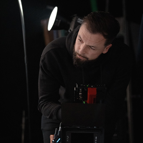 studio-video-rennes-cadreur-moniteur-camera Un cadreur professionnel barbu, vêtu d'un sweat à capuche noir, observe attentivement le moniteur de contrôle de sa caméra lors d'un tournage au Studio 401 à Rennes. La scène, capturée dans la pénombre du plateau avec un projecteur de studio en arrière-plan, souligne la concentration et l'expertise technique lors d'une production audiovisuelle.