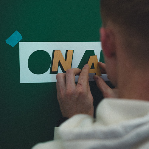 studio-video-rennes-construction-decor-on-air Vue en gros plan de dos d'un membre de l'équipe du Studio 401 à Rennes, manipulant avec précision des lettres en bois pour composer l'inscription "ON AIR" sur un mur vert foncé. Un pochoir blanc et du ruban adhésif bleu servent de guides pour le positionnement, illustrant le travail de préparation manuelle et la personnalisation des décors pour les plateaux de tournage vidéo et les studios de podcast.
