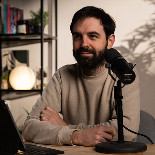 studio-video-rennes-enregistrement-podcast-intervenant Un homme barbu portant un sweat beige est assis à une table en bois devant un microphone professionnel Shure SM7B lors d'un tournage au Studio 401 à Rennes. Le décor en arrière-plan est composé d'une étagère noire avec des livres, une lampe sphérique lumineuse et des plantes vertes, créant une ambiance feutrée et qualitative pour un podcast ou une interview vidéo. Un clavier de tablette est partiellement visible au premier plan.