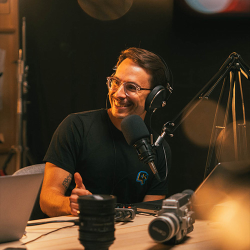 studio-video-rennes-enregistrement-podcast-micro-professionnel Un homme souriant portant des lunettes et un casque audio professionnel lors d'une session d'enregistrement de podcast au Studio 401 à Rennes. Il est installé devant un microphone de studio de haute qualité monté sur un bras articulé. Le décor inclut une table en bois avec du matériel technique, notamment des ordinateurs portables et des optiques de caméra, créant une atmosphère de production audiovisuelle moderne et conviviale.