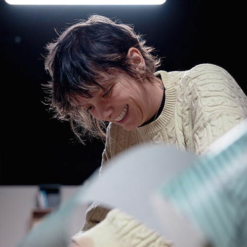 studio-video-rennes-equipe-preparation-decor-sourire Une femme aux cheveux courts et bruns, portant un pull torsadé beige, sourit en travaillant sur la fabrication d'un élément de décor au Studio 401 à Rennes. La prise de vue en contre-plongée avec un éclairage de studio souligne l'aspect artisanal et l'énergie positive de l'équipe technique lors de la préparation des plateaux pour un tournage vidéo.