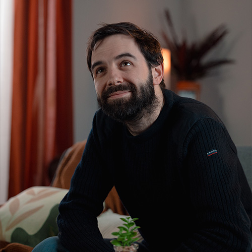 studio-video-rennes-location-decor-appartement-homme-01 Portrait d'un homme barbu en pull noir, souriant et regardant vers le haut, assis dans le décor "appartement" du Studio 401 à Rennes. L'arrière-plan flou montre des rideaux orange, des coussins à motifs et une ambiance de salon chaleureuse, illustrant la qualité des plateaux aménagés pour le tournage d'interviews ou de fictions.