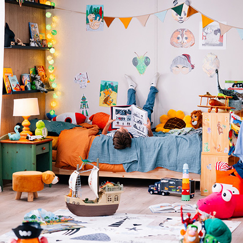 studio-video-rennes-location-decor-chambre-enfant-04 Un décor de chambre d'enfant richement décoré au Studio 401 à Rennes, conçu pour des productions vidéo et shootings photos. L'image montre un enfant allongé sur un lit, les pieds contre le mur, lisant un livre dans un univers coloré rempli de jouets, de dessins de dinosaures, de peluches et de guirlandes, illustrant la capacité du studio à créer des environnements domestiques réalistes et chaleureux.