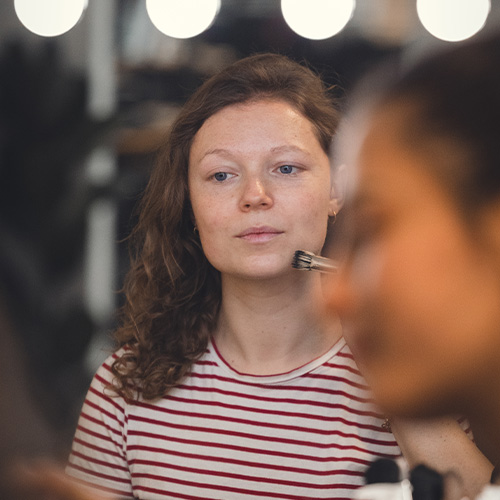 studio-video-rennes-maquillage-hmc-loges-01 Une jeune femme aux cheveux bouclés, vêtue d'un t-shirt rayé rouge et blanc, se fait maquiller le visage à l'aide d'un pinceau professionnel dans l'espace loges du Studio 401 à Rennes. En arrière-plan, des ampoules de miroir de loge créent des cercles de lumière flous (bokeh), soulignant l'atmosphère de préparation HMC (Habillage, Maquillage, Coiffure) avant un tournage vidéo ou une séance photo.