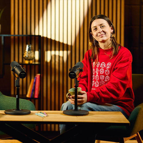studio-video-rennes-plateau-podcast-decor-bois-02 Une femme portant un sweat rouge et tenant une tasse est installée sur un plateau de podcast au Studio 401 à Rennes. Le décor se compose de fauteuils scandinaves verts, de deux micros professionnels Shure SM7B sur une table en bois, et d'un arrière-plan élégant avec des tasseaux de bois, une étagère décorative et un tube LED vertical, créant une ambiance chaleureuse pour le tournage d'émissions ou d'interviews.