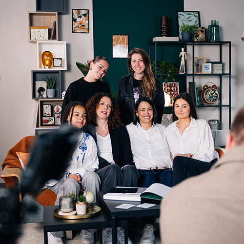 studio-video-rennes-tournage-equipe-decor-salon-02 Un groupe de six personnes pose avec le sourire sur un canapé gris dans le décor salon du Studio 401 à Rennes. Au premier plan, on aperçoit de dos un cadreur et le moniteur de sa caméra capturant la scène. Le plateau est soigneusement aménagé avec un mur de fond vert canard, des étagères décoratives modernes et un éclairage professionnel, illustrant les possibilités de mise en scène pour des interviews ou des fictions.