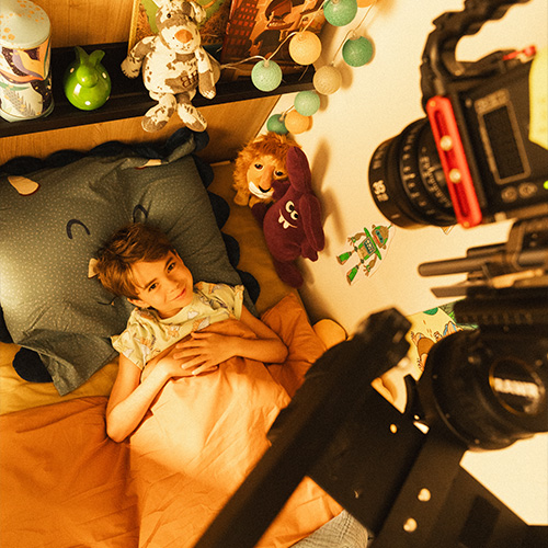 studio-video-rennes-tournage-fiction-decor-chambre-enfant-01 Un jeune garçon allongé dans un lit sur un plateau de tournage au Studio 401 à Rennes, recréant l'ambiance d'une chambre d'enfant. Le décor est agrémenté de peluches, d'une guirlande lumineuse et de livres colorés. Une caméra professionnelle, positionnée en plongée au-dessus du lit, capture la scène, illustrant l'expertise du studio dans la création de décors réalistes pour des projets de fiction ou de publicité.