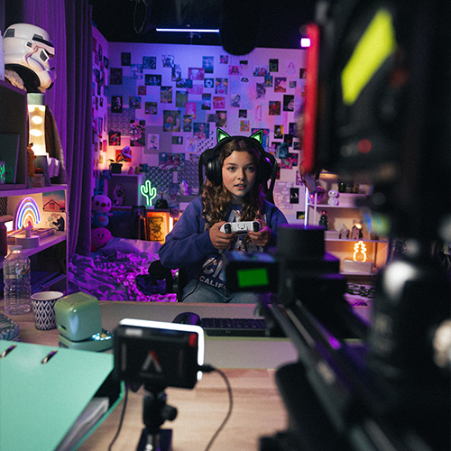 studio-video-rennes-tournage-gaming-decor-streaming-01 Une jeune fille portant un casque audio avec des oreilles de chat lumineuses est filmée sur un plateau thématique au Studio 401 à Rennes. Elle tient une manette de jeu vidéo dans un décor de style "gaming/streaming" immersif, composé de nombreux néons (cactus, arc-en-ciel, ananas), de posters muraux et d'accessoires de pop culture. La prise de vue inclut le matériel de tournage au premier plan (caméra et moniteur), mettant en avant l'aspect professionnel de cette production réalisée dans un environnement richement décoré et coloré.