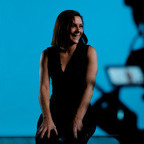 studio-video-rennes-tournage-interview-fond-bleu-01 Une femme souriante assise sur le plateau du Studio 401 à Rennes lors d'un tournage d'interview professionnelle. Elle est mise en valeur par un éclairage de studio précis sur un fond bleu uni (cyclorama). Au premier plan, la silhouette floue d'une caméra et d'un moniteur de contrôle souligne l'environnement de production audiovisuelle et la qualité technique des captations réalisées dans cet espace de tournage.