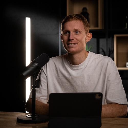 studio-video-rennes-tournage-podcast-micro-shure Un homme en t-shirt blanc s'exprime devant un microphone professionnel Shure SM7B lors d'un enregistrement de podcast au Studio 401 à Rennes. Il est installé à une table en bois avec une tablette numérique devant lui. Le décor d'arrière-plan est sombre et élégant, mis en valeur par un tube LED vertical blanc et des étagères décoratives, offrant un cadre moderne pour la production de contenus digitaux.